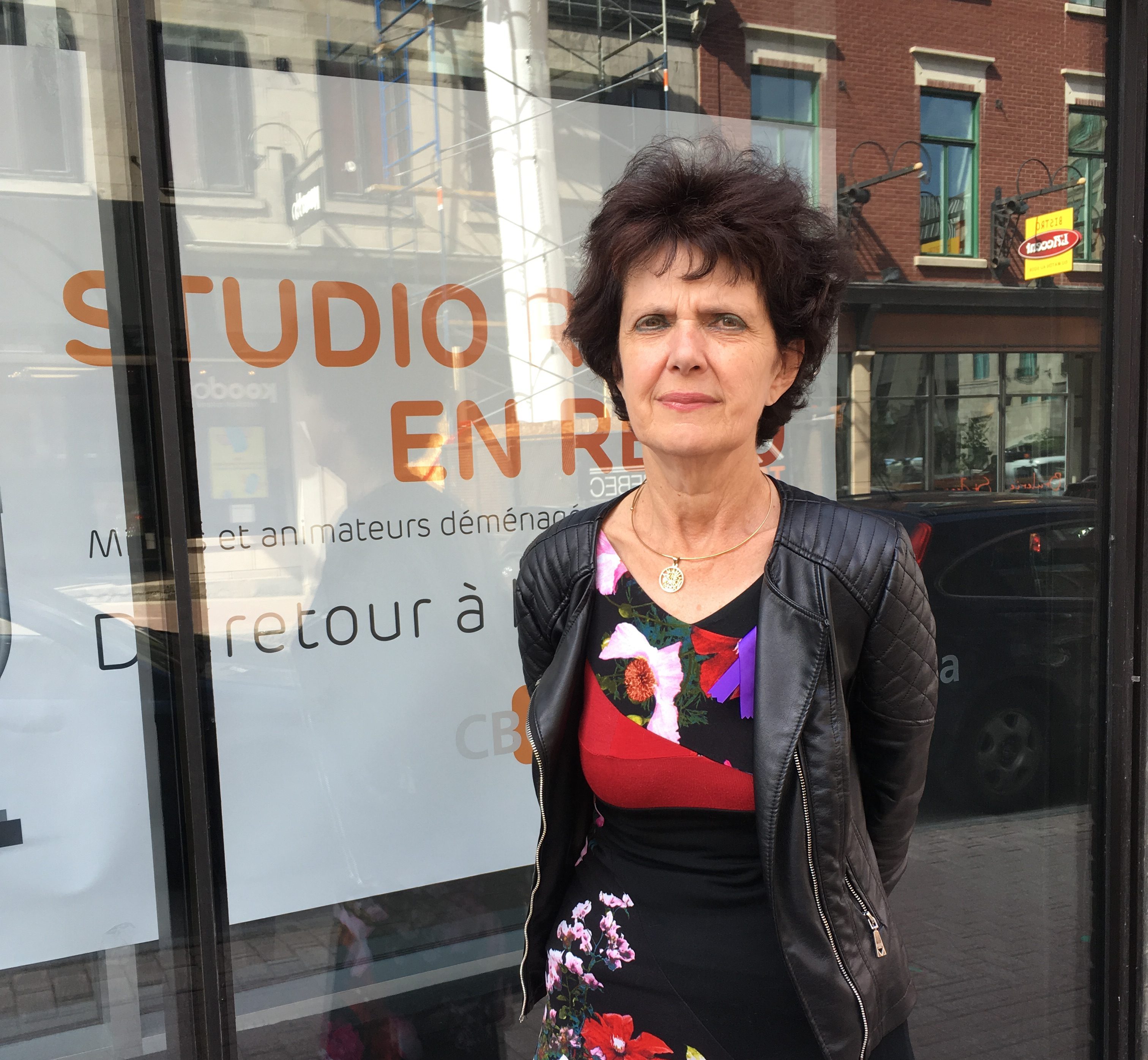 Photo de madame Gagnon, cheveux semi-long brun, veston de cuir et robe à motifs modernes rouge et noir, devant la fenêtre d'un studio.